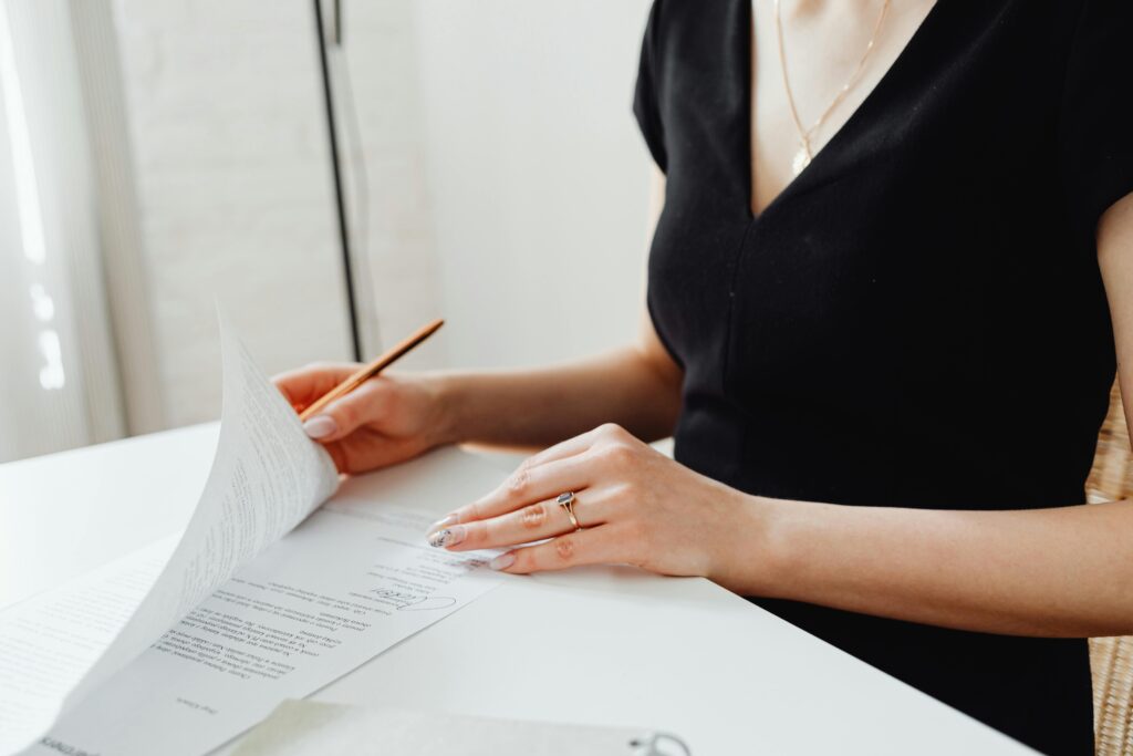 A woman in business attire reviewing and signing a legal document at a desk. She holds a pen while flipping through paperwork