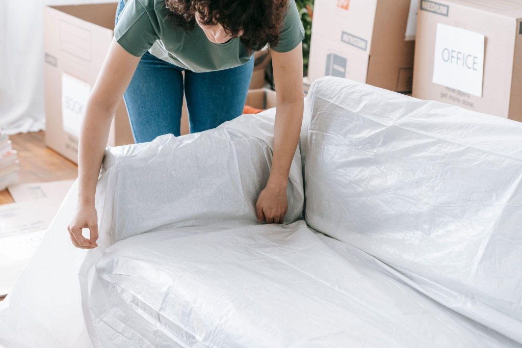 Mover wrapping a sofa in protective plastic beside labeled moving boxes.